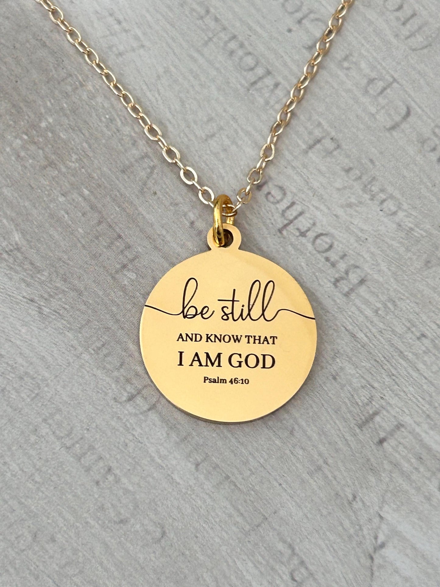 Gold-colored pendant necklace with a round charm. The charm features the words "be still" above the phrase "AND KNOW THAT I AM GOD" and "Psalm 46:10" in a black, elegant font. The necklace has a delicate chain.