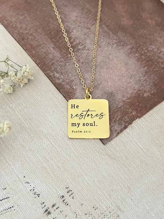 May include: A gold necklace with a square pendant that says 'He restores my soul. Psalm 23:3'.