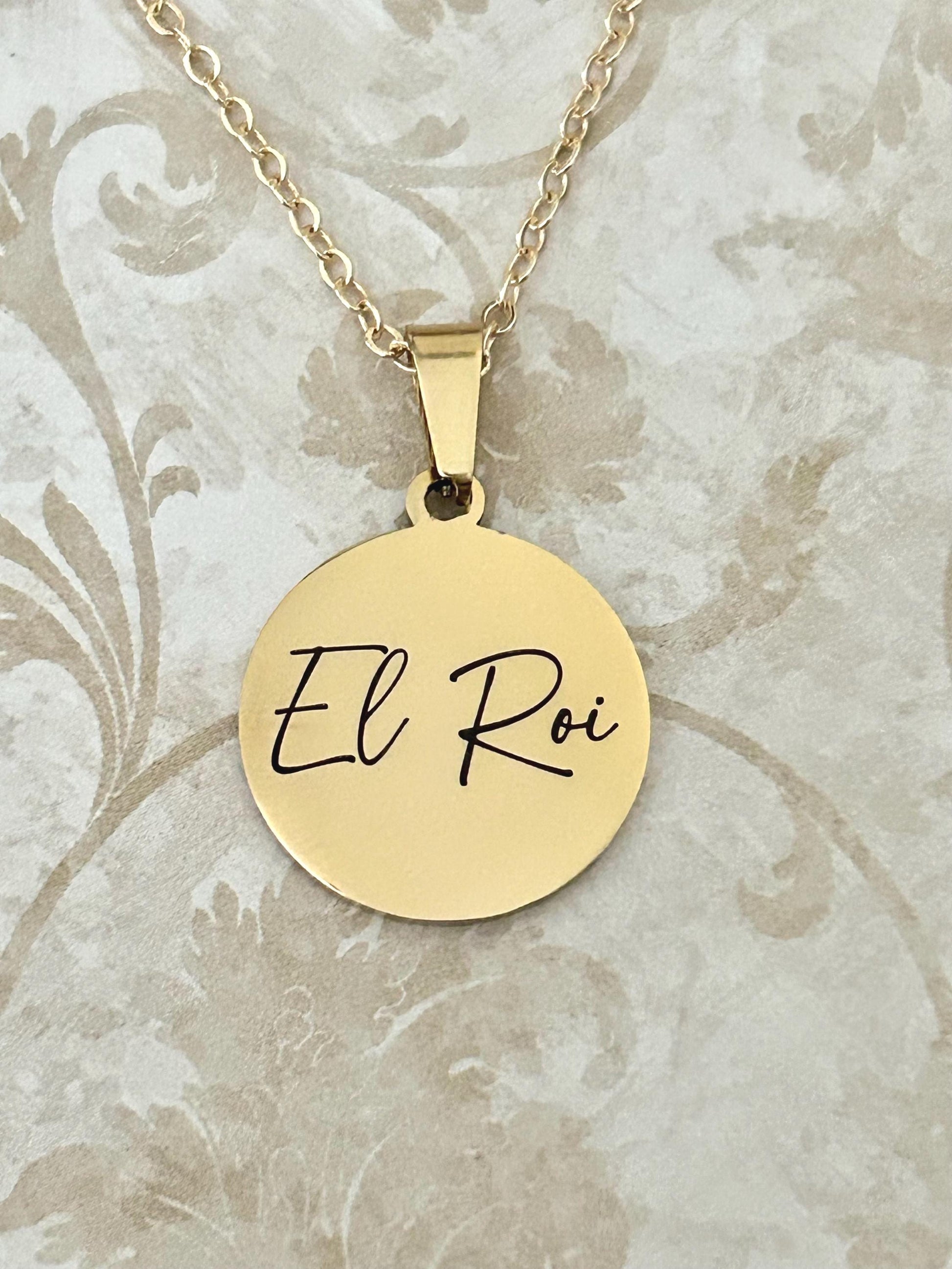 A gold-colored necklace with a circular pendant. The pendant has the words "El Roi" in black script. The necklace chain is also gold-colored and delicate. The background is a floral pattern in neutral tones.