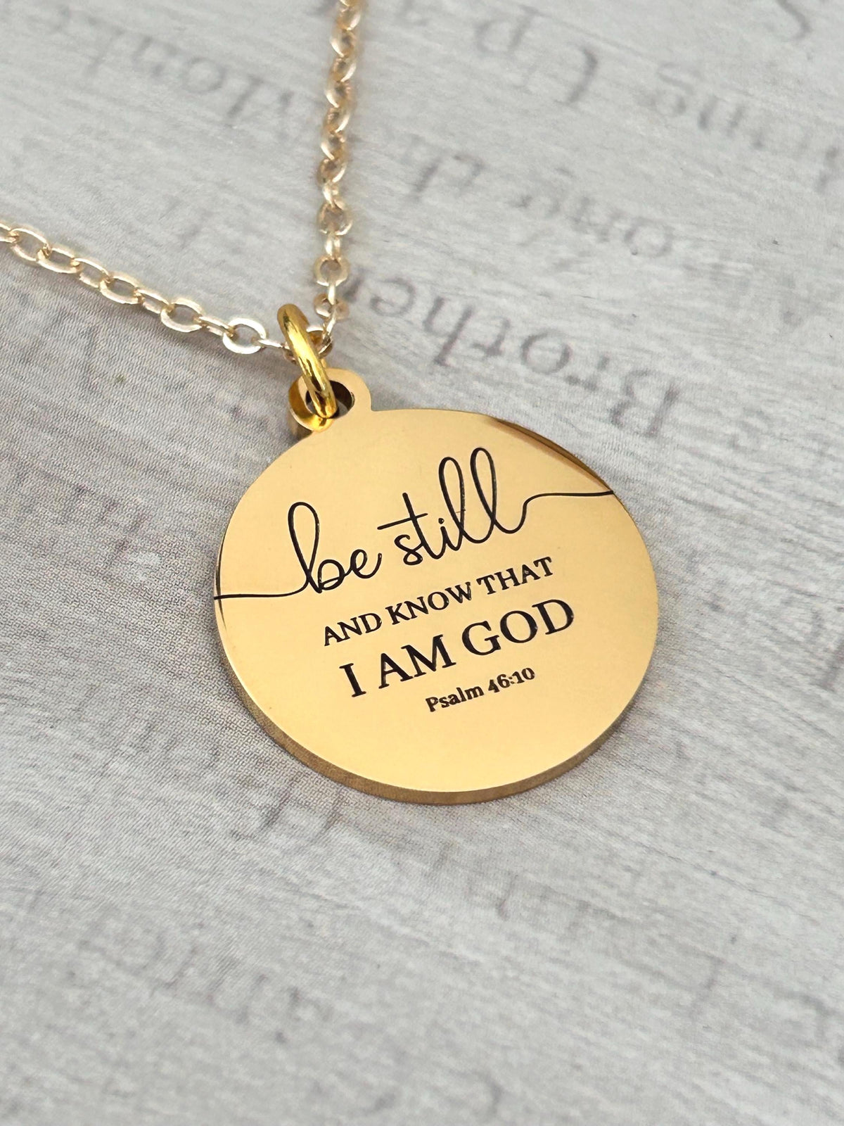 A gold-colored pendant necklace with the inscription "be still AND KNOW THAT I AM GOD Psalm 46:10" in black lettering. The pendant is circular and hangs from a delicate gold chain.