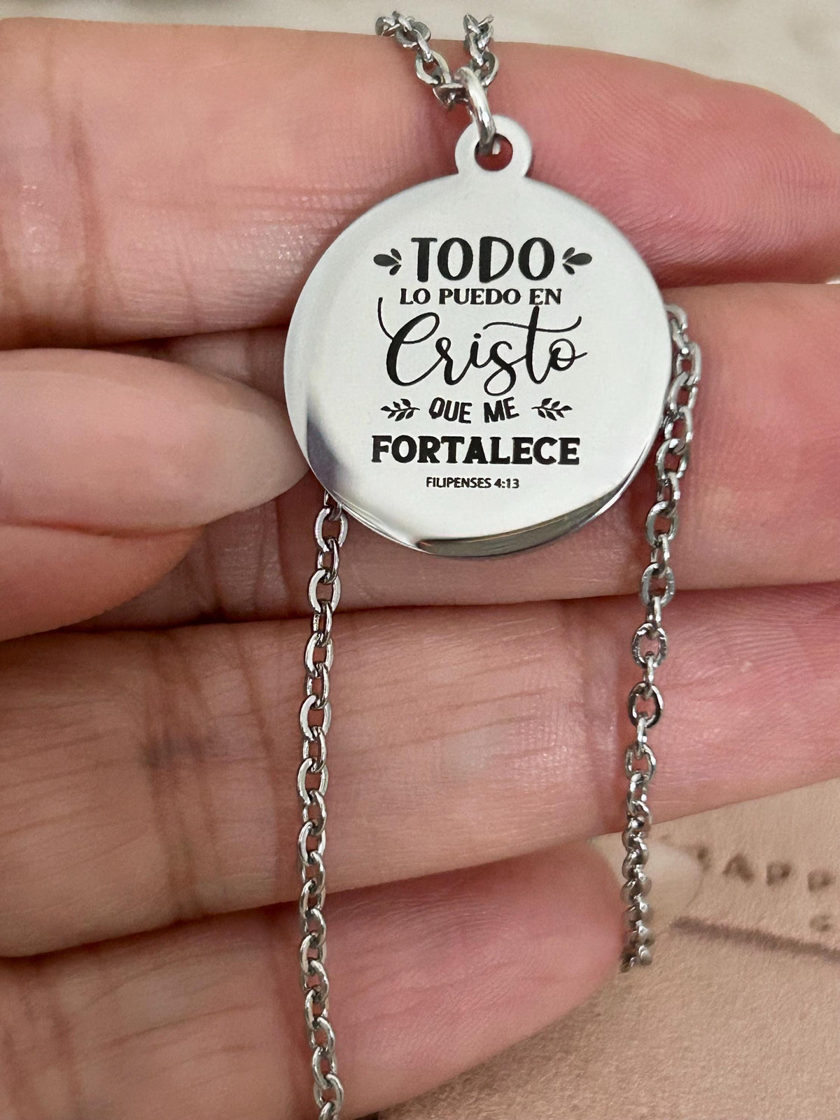 May include: A silver-colored necklace featuring a circular pendant with the engraved text "TODO LO PUEDO EN CRISTO QUE ME FORTALECE" and "FILIPENSES 4:13". The pendant hangs from a delicate chain.