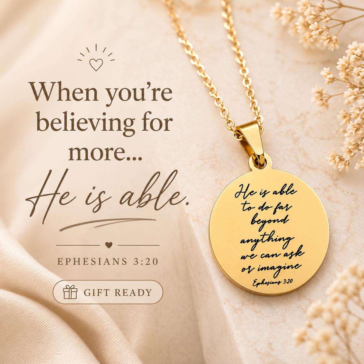 May include: A gold-colored pendant necklace with a round charm. The charm has an engraved message and the text "He is able to do far beyond anything we can ask or imagine". The necklace is displayed on a soft, beige background.